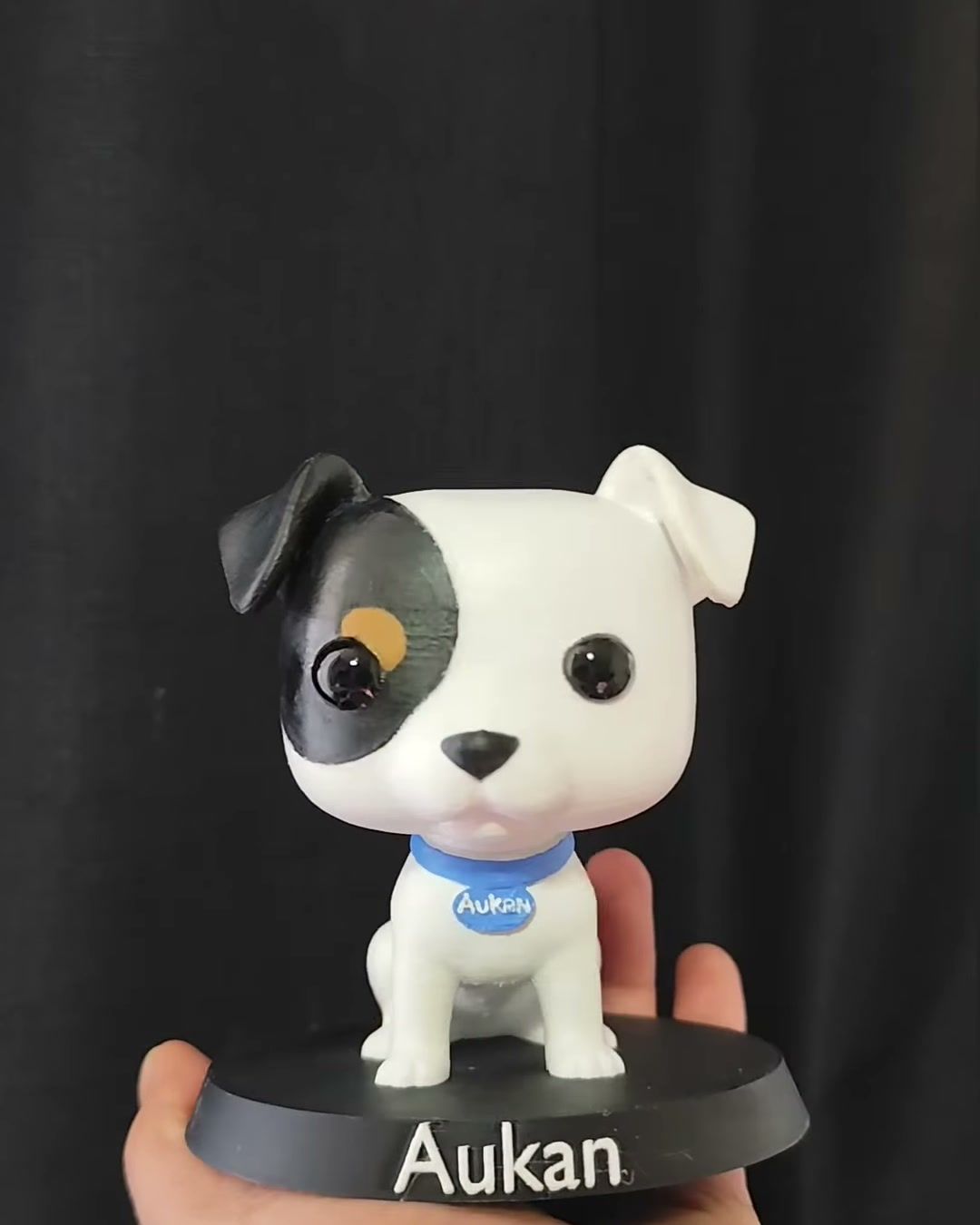 Figura de perro blanco con una mancha negra en el ojo y collar azul, sobre base negra con el nombre Aukan.