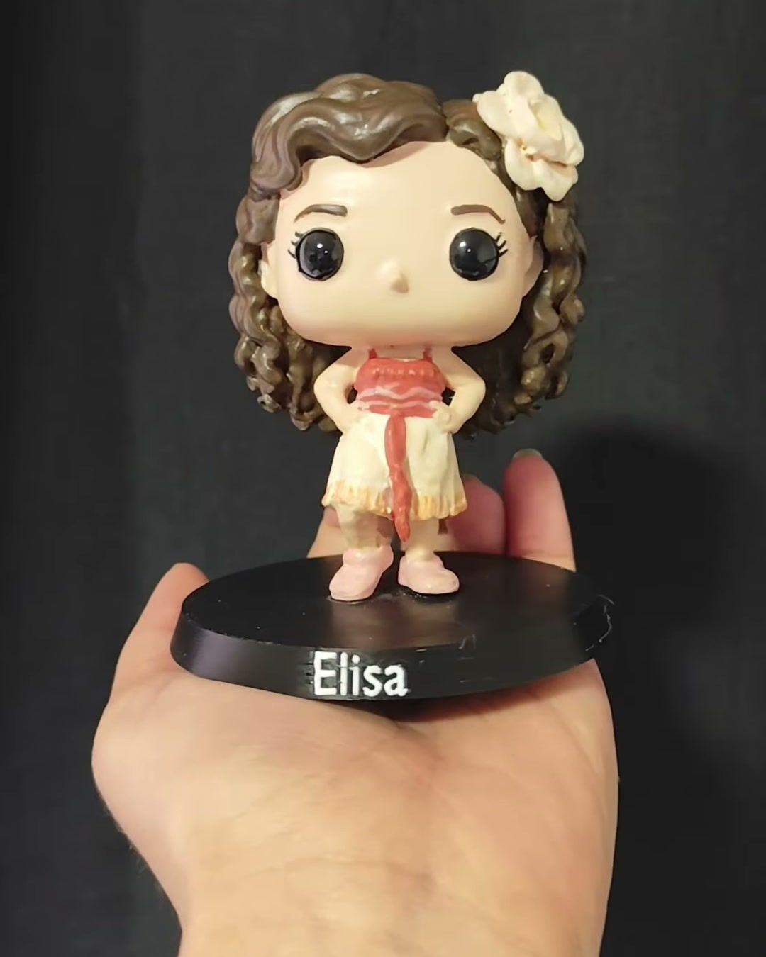 Figura de una niña de pelo castaño ondulado con una flor blanca, vestido claro y cinta roja, sobre base negra con el nombre Elisa.