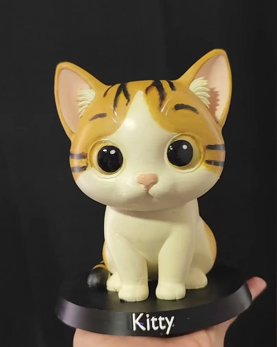 Figura de gata atigrada crema y café de ojos amarillos, sobre base negra con el nombre Kitty.