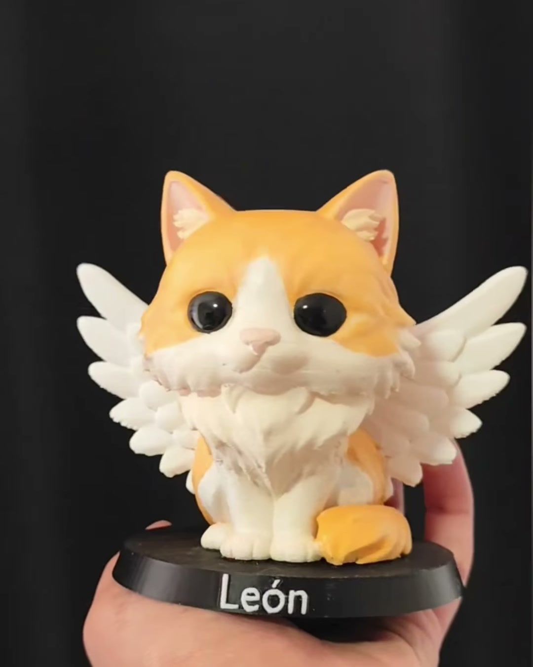 Figura de gato naranjo y blanco de pelo largo con alas de ángel blancas, sobre base negra con el nombre León.