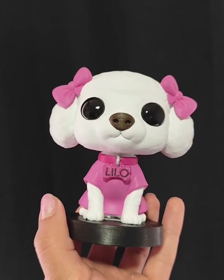 Figura de perrita blanca con moños rosados y polera rosada con su nombre, sobre base negra.