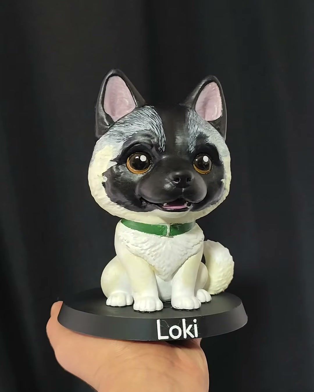 Figura de perro de pelaje gris, negro y blanco con collar verde, sobre base negra con el nombre Loki.