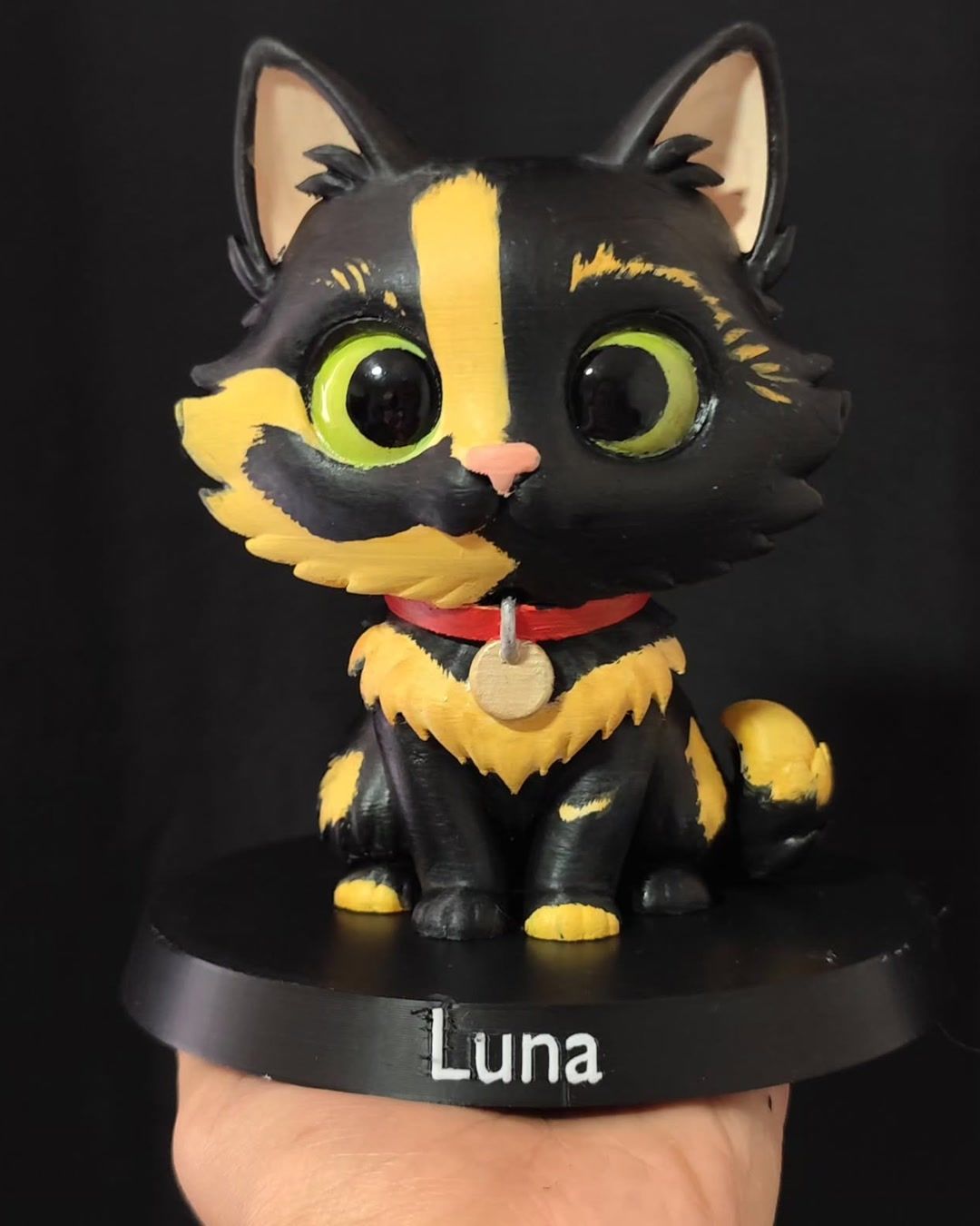 Figura de gata negra con manchas amarillas y ojos verdes, collar rojo con placa dorada, sobre base negra con el nombre Luna.