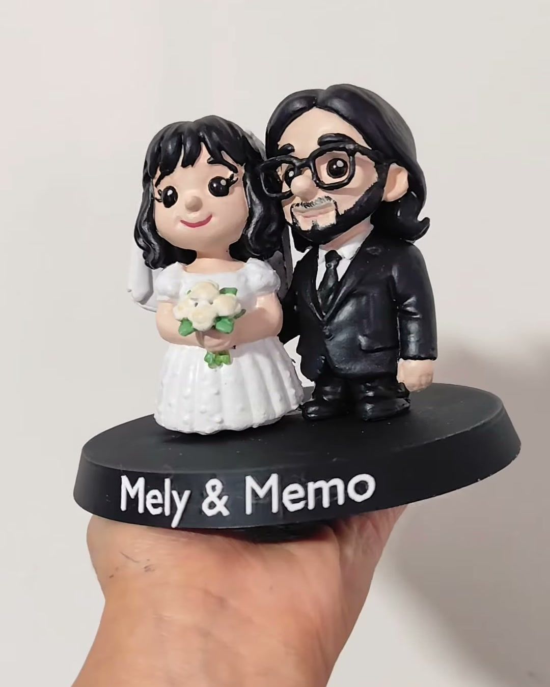 Figura de pareja de novios: ella con vestido blanco y ramo, él con traje negro, lentes y barba, sobre base negra con sus nombres.