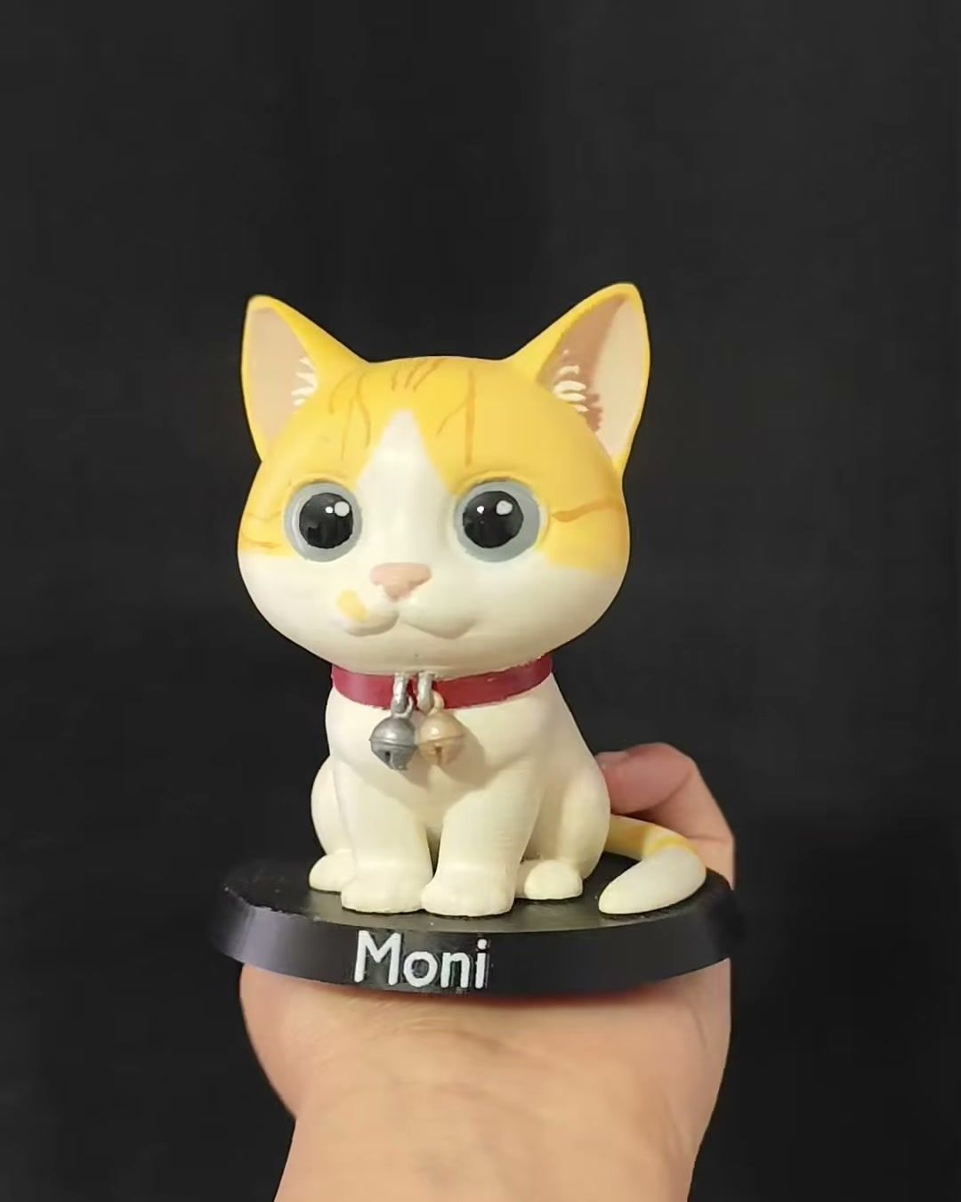 Figura de gata blanca con manchas naranjas y collar rojo con cascabel, sobre base negra con el nombre Moni.