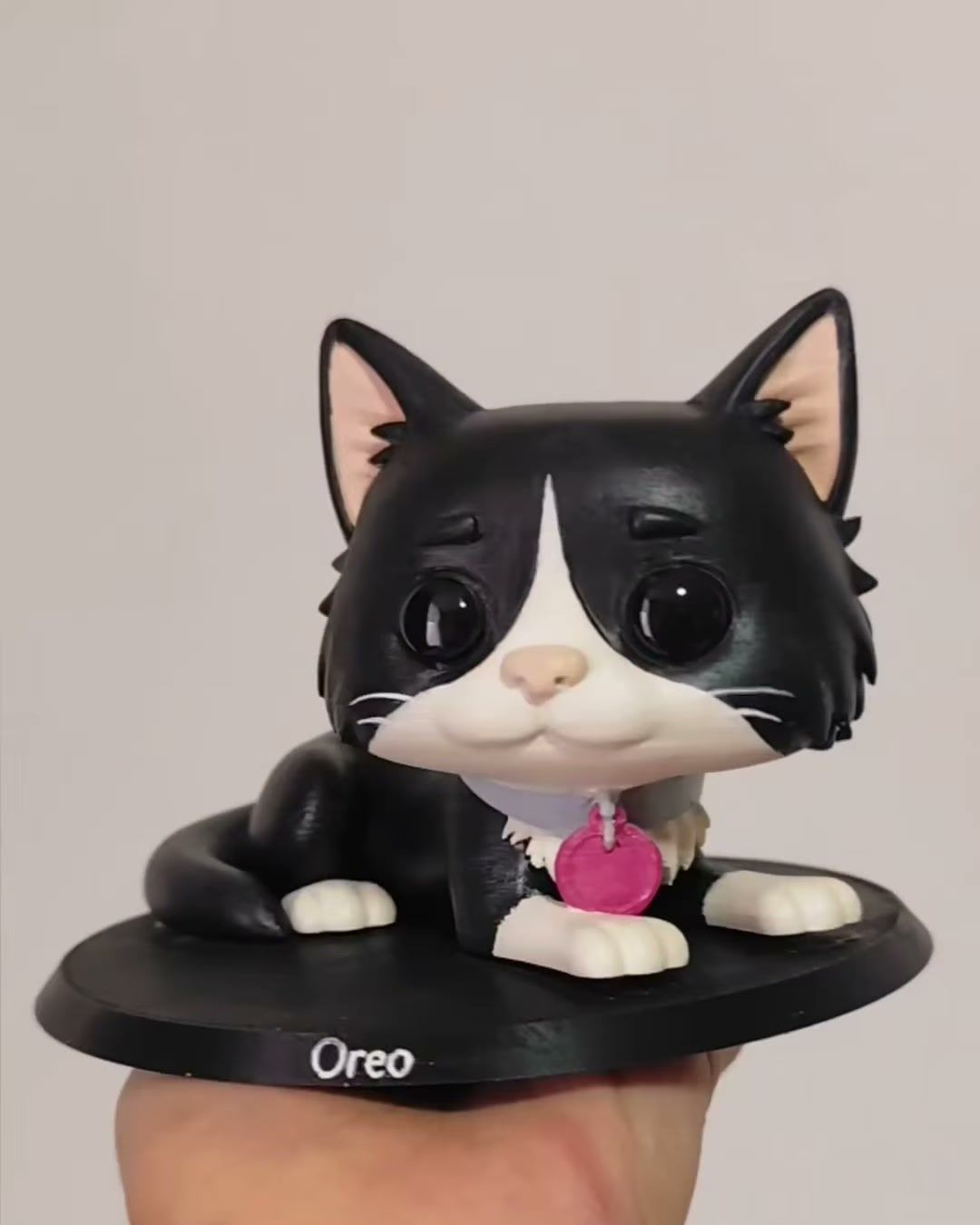 Figura de gato blanco y negro recostado, con placa rosada en el collar, sobre base negra con el nombre Oreo.