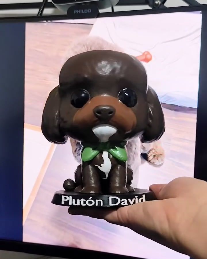 Figura de perro de pelo café oscuro con pañuelo verde, sostenida frente a la foto del perro real en una pantalla.