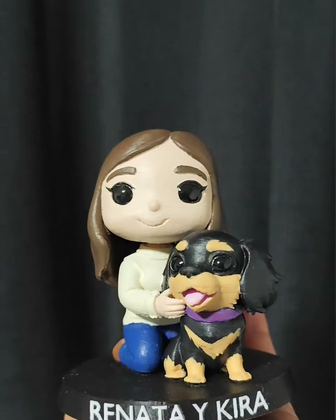 Figura de una mujer de pelo castaño abrazando a su perrita negra y café, sobre base negra con los nombres Renata y Kira.