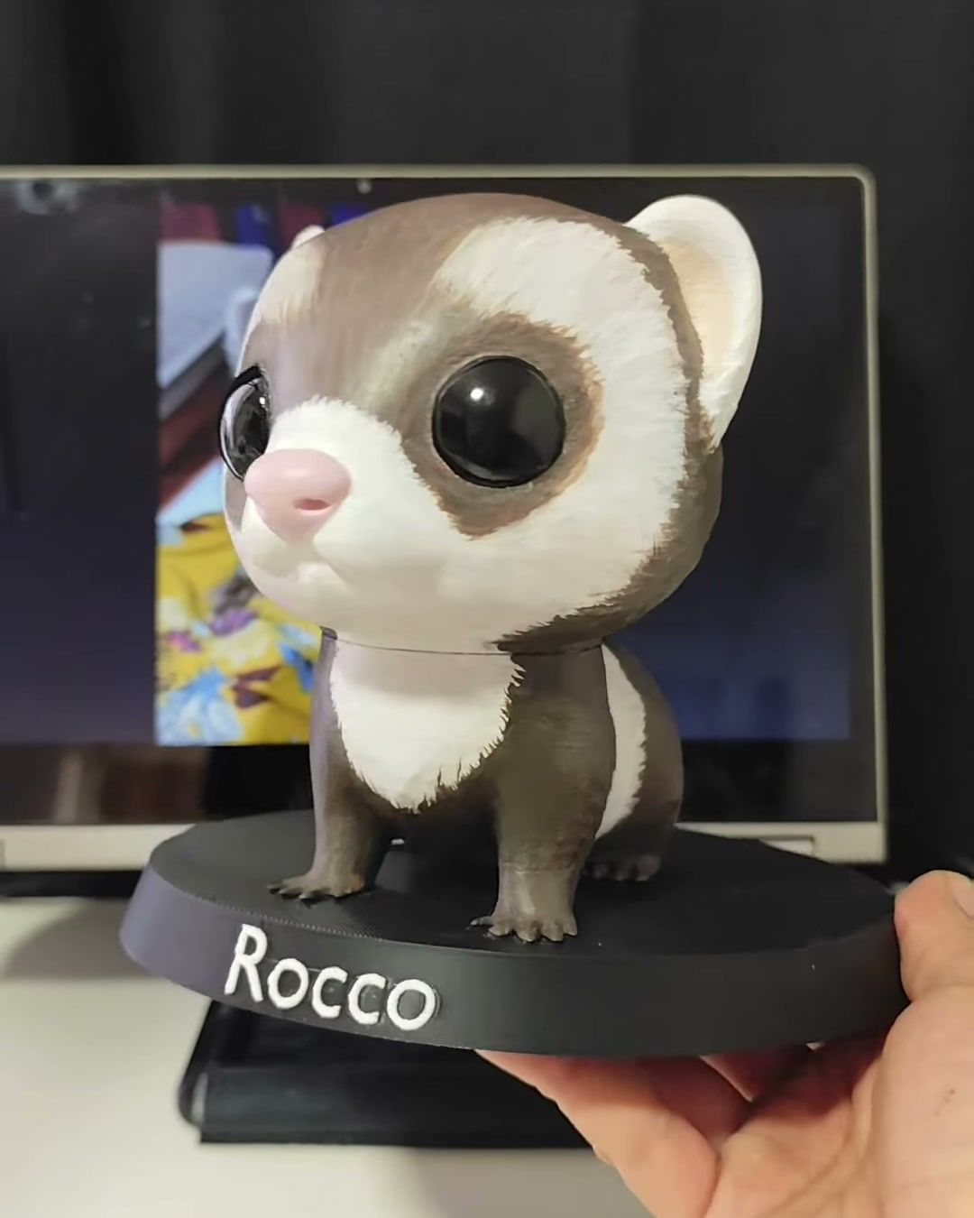Figura de hurón café y blanco de ojos grandes, sobre base negra con el nombre Rocco, frente a una pantalla.