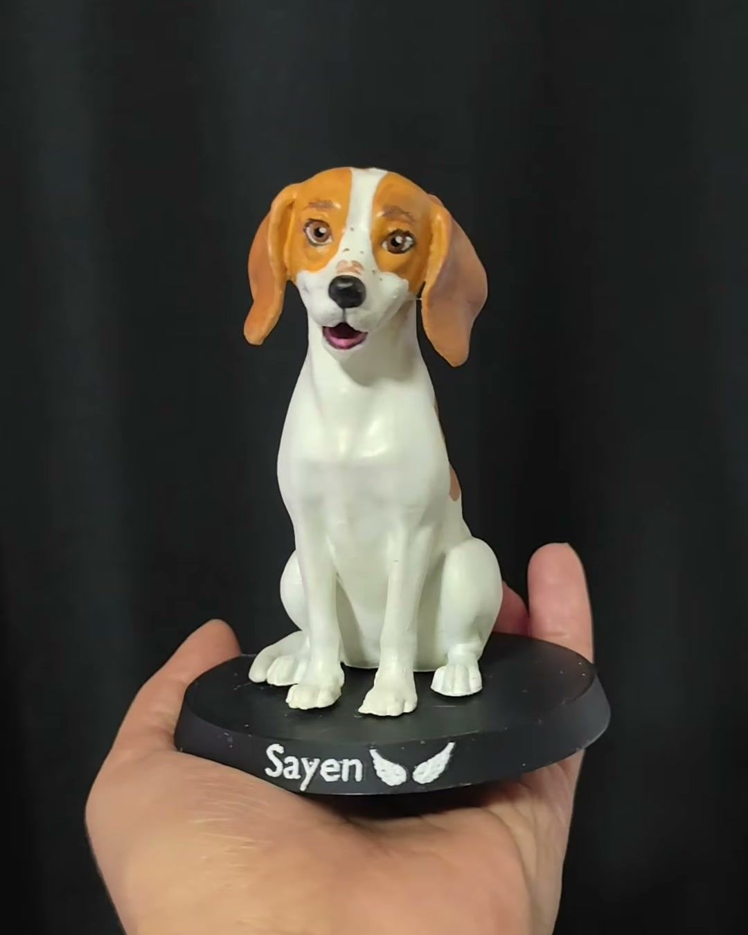 Figura de perra blanca con orejas y cara café, sentada sobre base negra con el nombre Sayen.