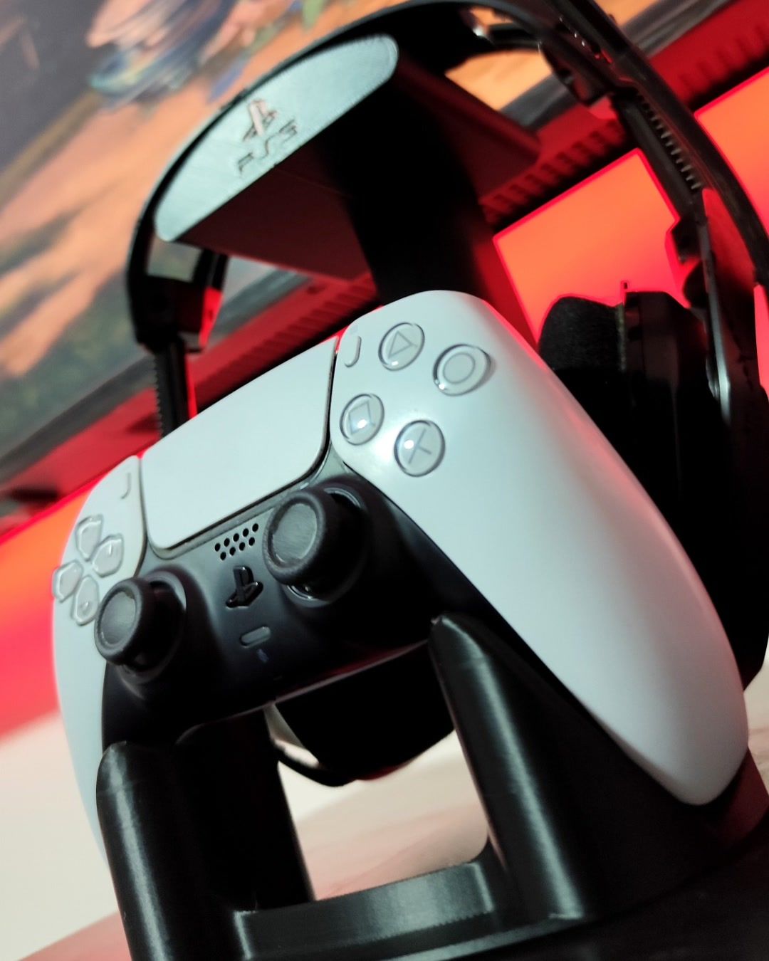 Soporte negro que sostiene un control de PlayStation 5 abajo y unos audífonos arriba, con luz roja de fondo.
